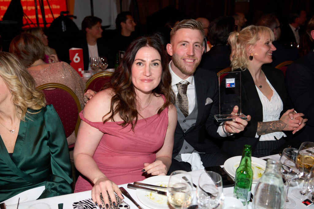 Rebecca Nilsson and Max Malm are sitting at the dinner table at the gala, holding the Great Place to Work award with smiles on their faces.