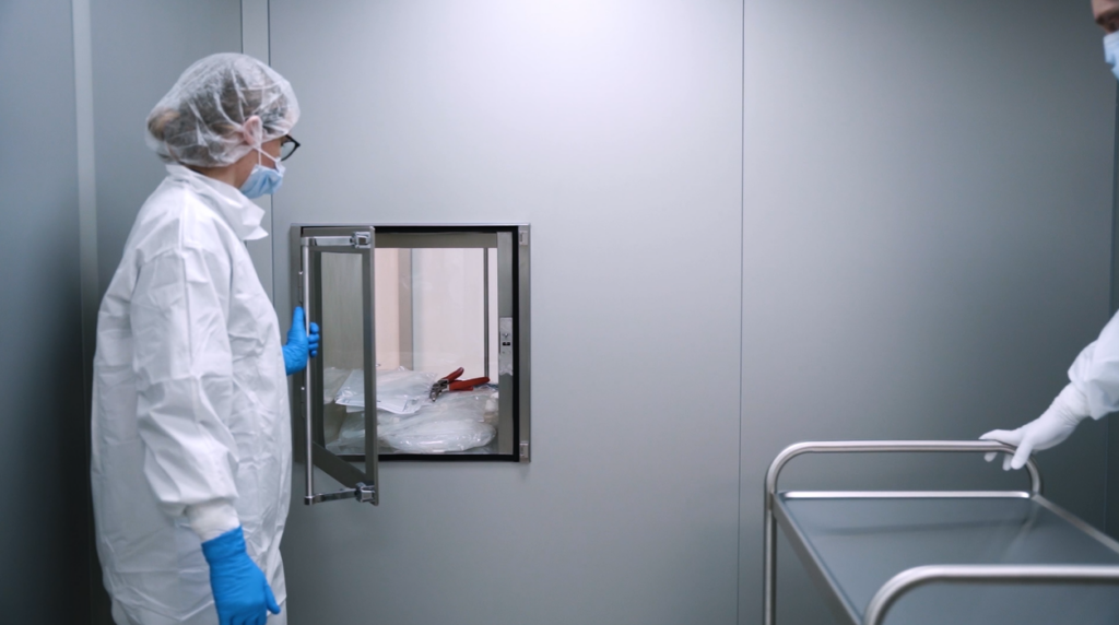 Två medarbetare i vita renrumsdräkter arbetar vid en luftsluss och hanterar material mellan olika ISO-klassade zoner i Colly Flowtechs renrum.